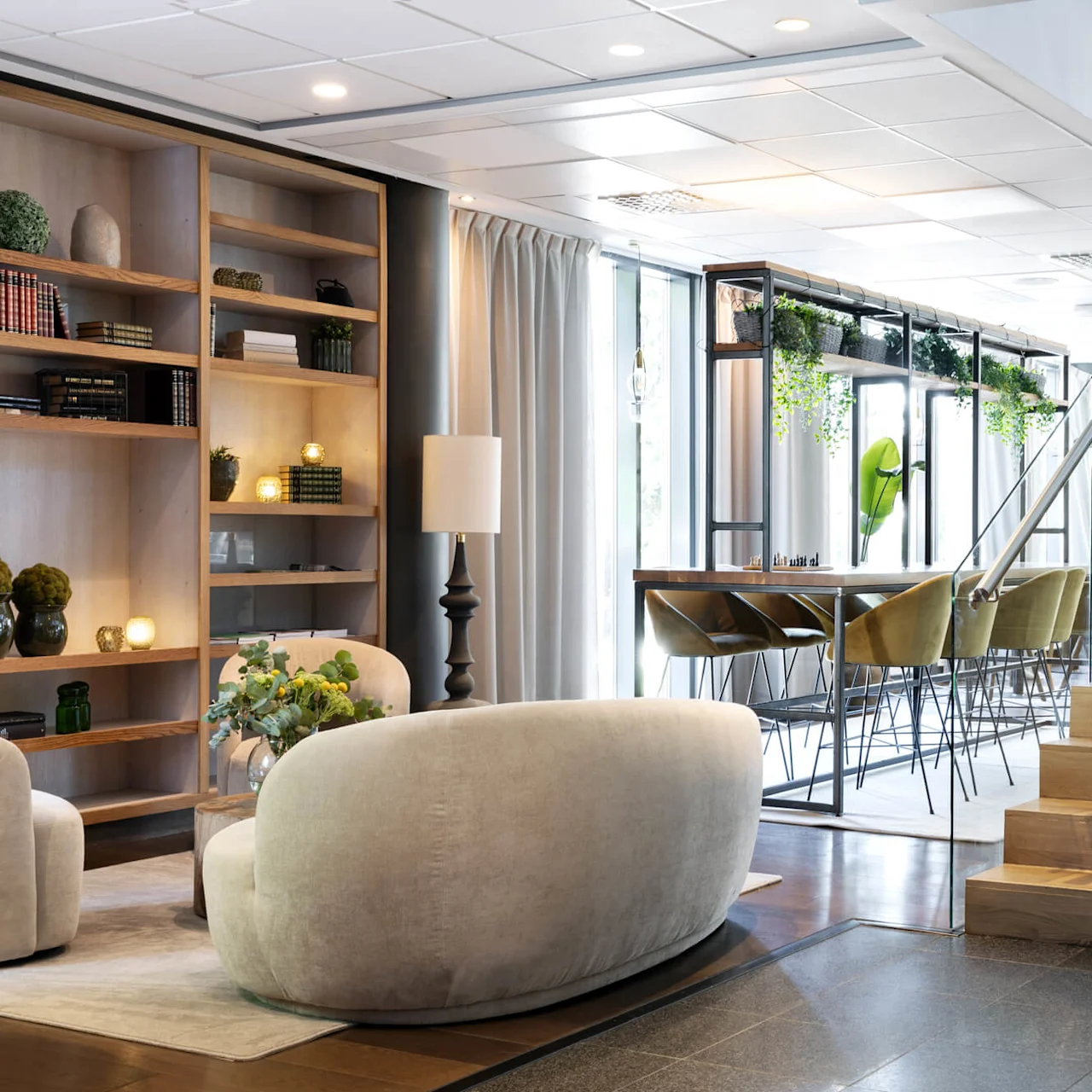 A spacious modern interior featuring a cozy seating area with plush sofas near wooden shelves. Adjacent is a dining table with green chairs, under vibrant hanging plants, next to a staircase.