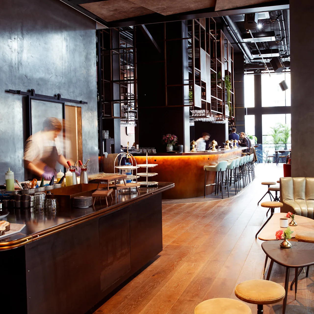 Chef prepares dishes in a modern, stylish restaurant with a curved bar and comfortable seating area, while patrons relax nearby.