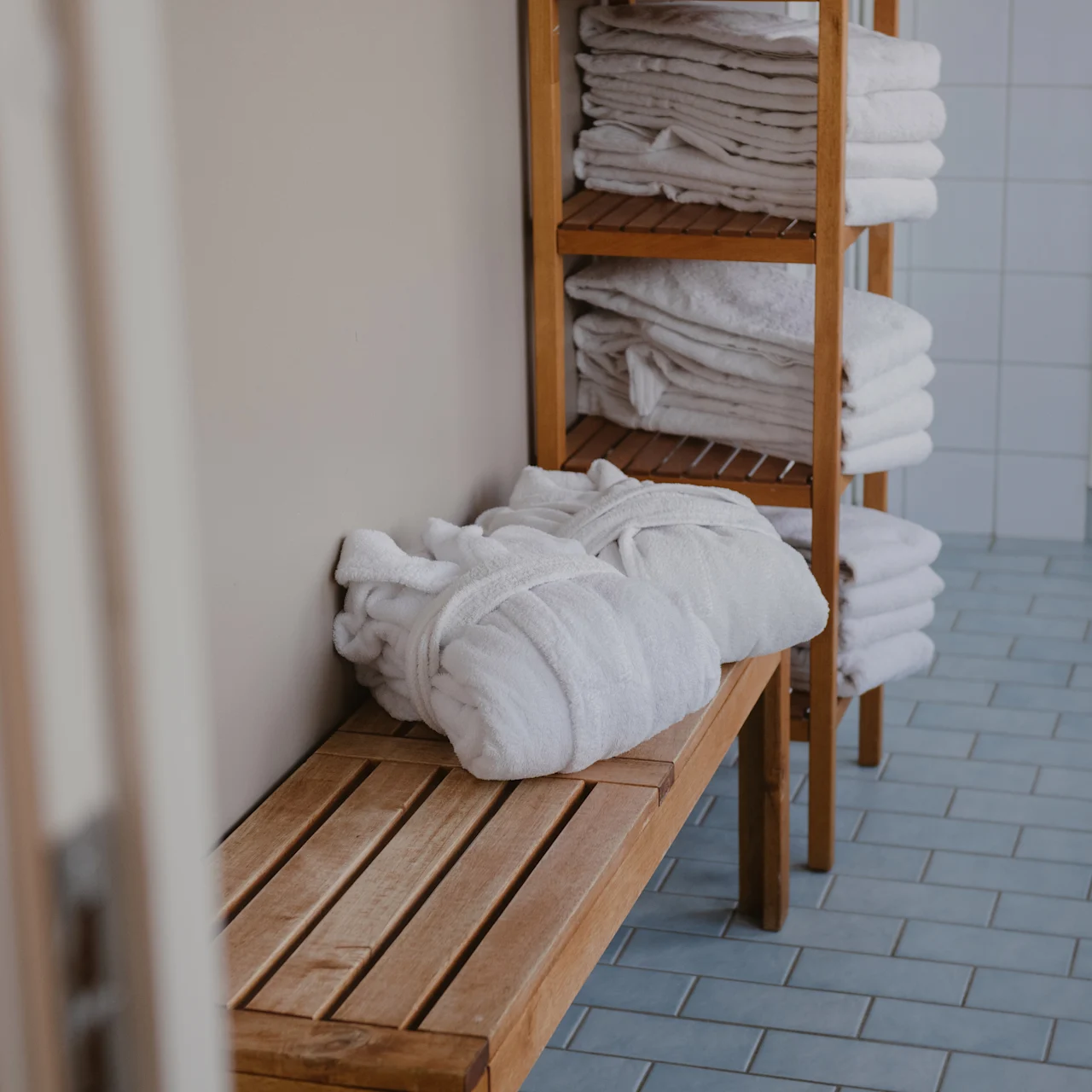 A white bathrobe rests on a wooden bench, surrounded by stacked towels on shelves in a tiled changing room.