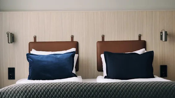 Two beds with brown leather headboards and blue pillows, covered by a quilted gray blanket in a modern, minimalistic room with light wood paneling and wall-mounted lamps.