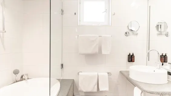 Ett ljust, modernt badrum med vita kakelplattor, med badkar, handfat och fräscha handdukar. Naturligt ljus kommer in genom ett fönster.