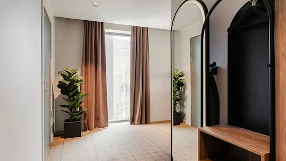 To grønne planter står i et moderne rum ved siden af gulvlange, brune gardiner med et spejlet garderobeskab og beige ternet tæppe under blødt lys.