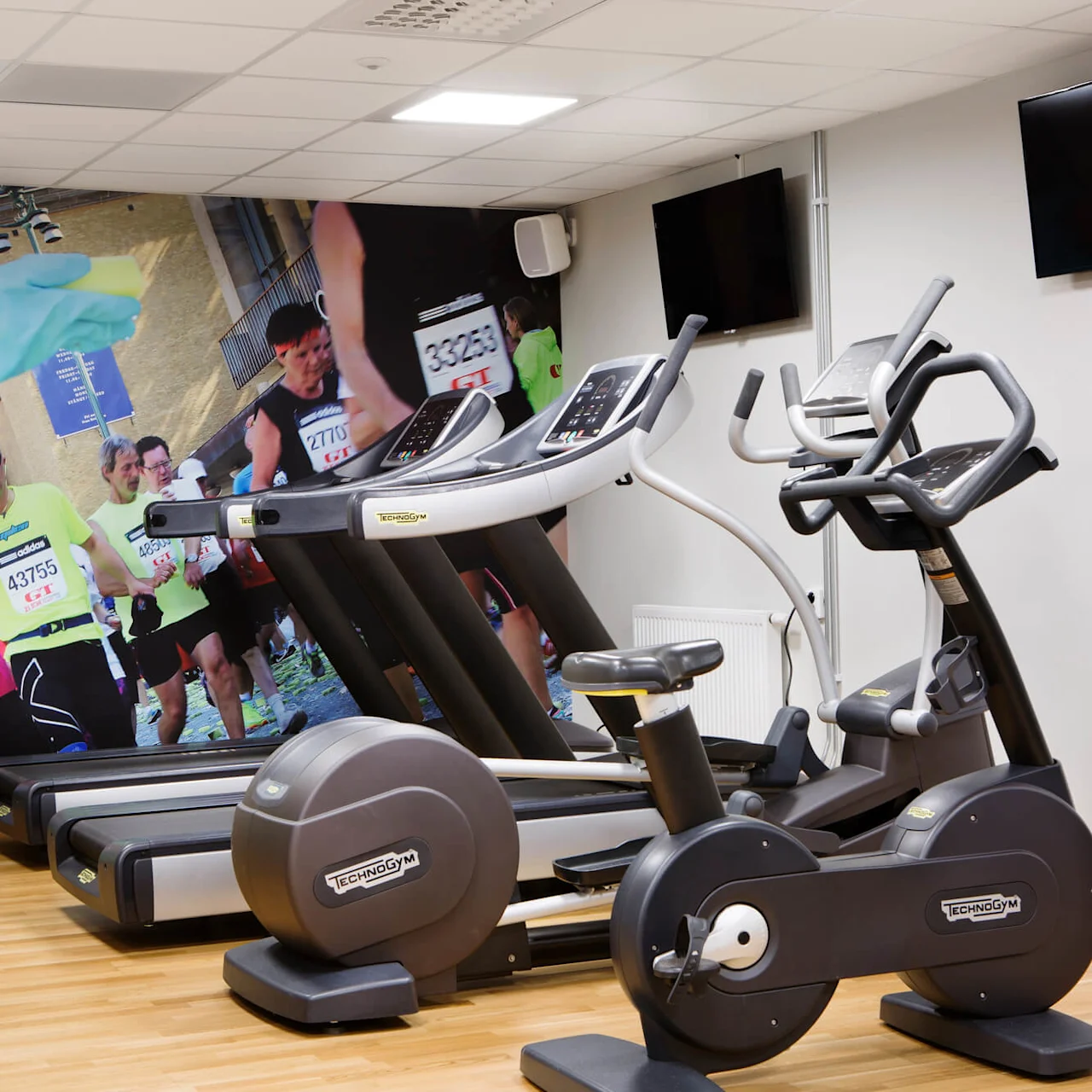 Löpband och träningscyklar står oanvända i ett gym. En väggmålning visar maratonlöpare i färgglada kläder. Två väggmonterade TV-apparater är avstängda.