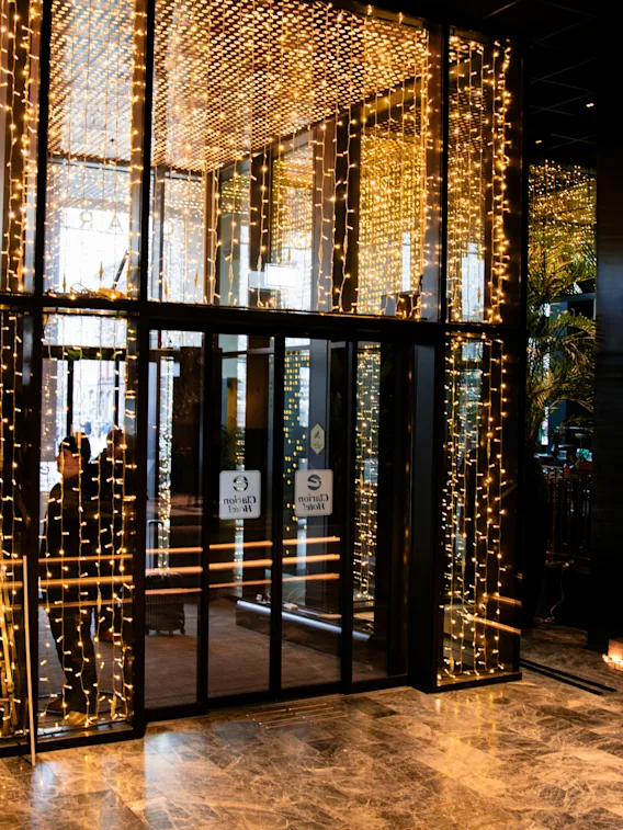 Glass doors adorned with warm string lights, creating a festive atmosphere in a hotel lobby. Reflections and marble flooring add elegance. Text on doors: “Clarion Hotel.”