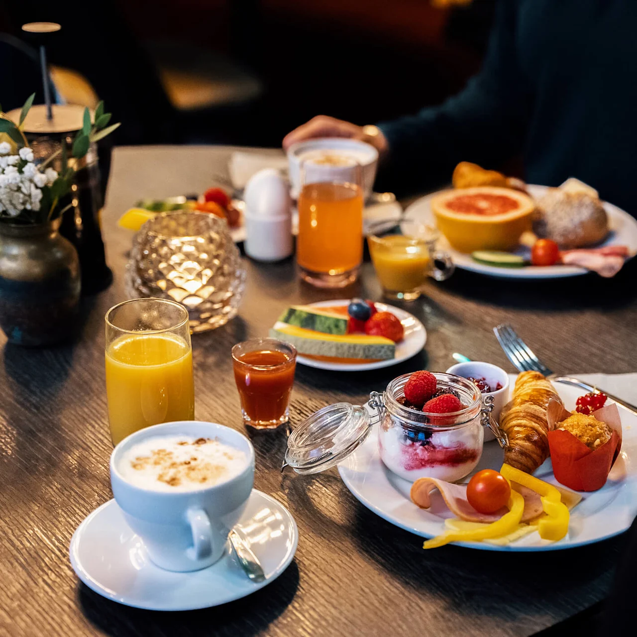 Frukost med cappuccino, apelsinjuice, frukt, yoghurtdessert, croissant, kallskuret och grönsaker på ett mörkt träbord.