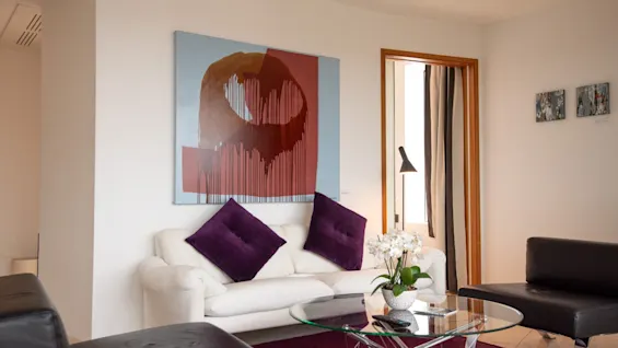 Ett modernt och inbjudande vardagsrum med en vit soffa, lila accenter, ett soffbord i glas och abstrakt konst på väggen.