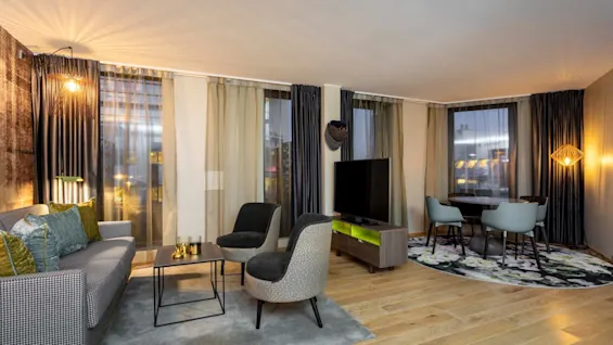 A living room with a grey sofa and armchairs surrounds a coffee table. A TV stands nearby. Dining area in the background. Wooden floor, large windows, and modern lighting.
