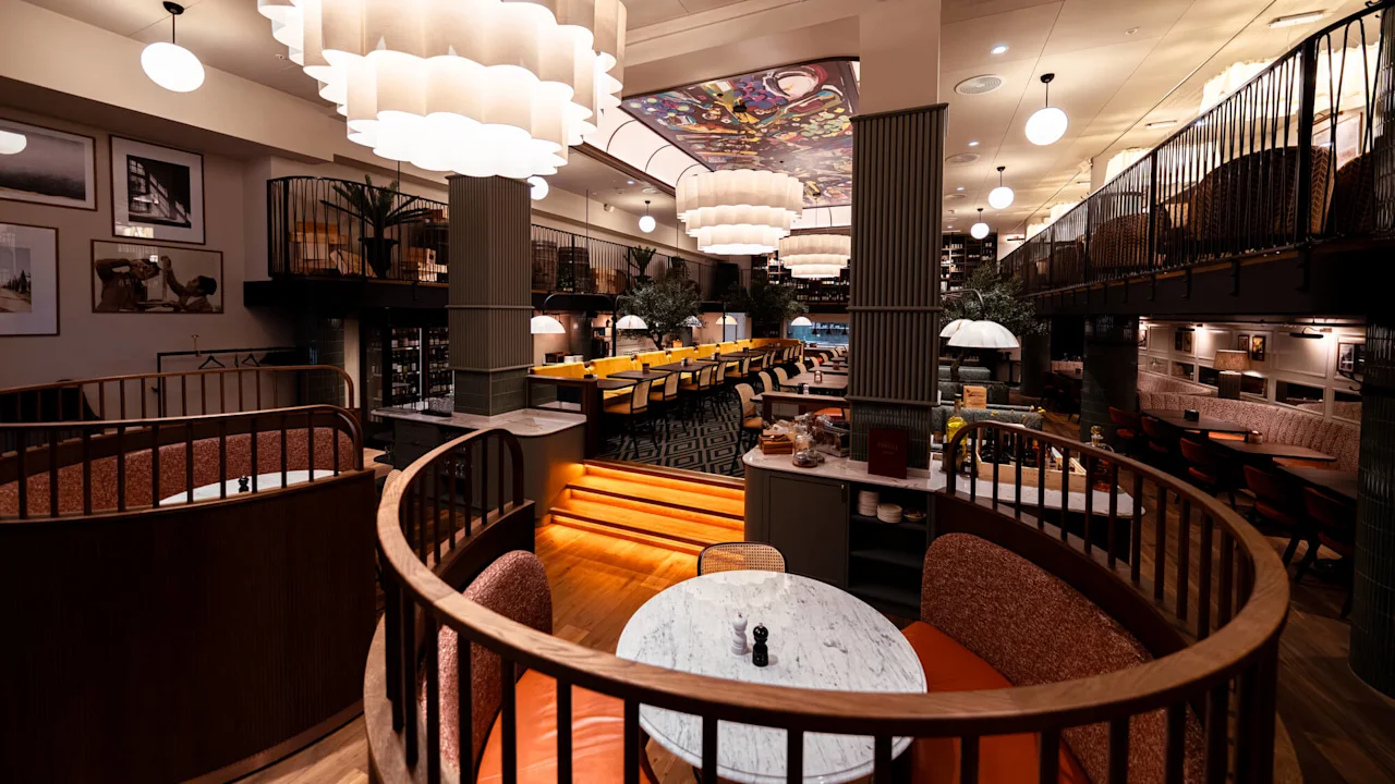 En restaurantinteriør med runde bokse og en central trappe. Over hænger store, bølgende pendellamper. De omkringliggende vægge viser indrammet kunstværk, hvilket bidrager til en hyggelig atmosfære.