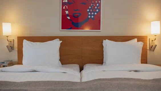 A neatly made double bed with white pillows and a gray blanket, flanked by wall-mounted lamps, beneath a colorful pop art portrait on the wall.