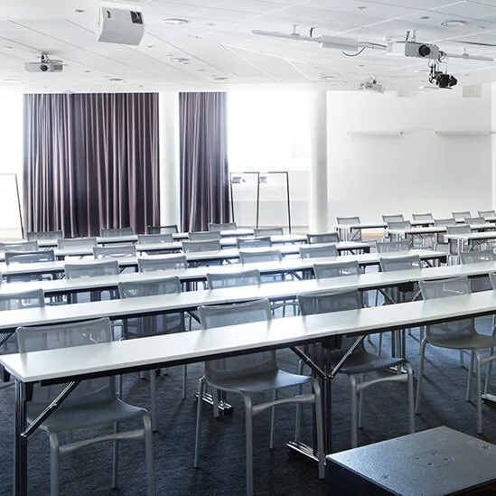 An empty conference room with rows of tables and chairs. A podium stands at the forefront. Ceiling lights and projectors are mounted above. Large curtains cover windows along one side.