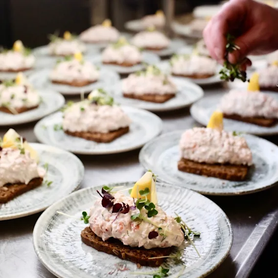 Swedish: Smörrebröd med krämig topping och citronklyfta på tallrikar. Hand garnerar med örter i ett kök.