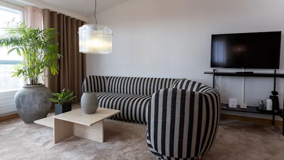 A striped black-and-white couch sits beside a wooden table with vases; a pendant light hangs above, and a potted plant rests near floor-length curtains. A wall-mounted TV completes the living room.