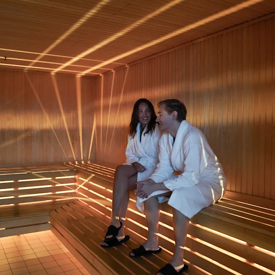 A man and a woman in white bathrobes sitting and relaxing inside a warm wooden sauna with indirect lighting.