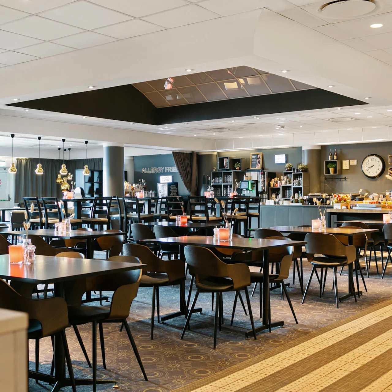 En rummelig spisesal er sat op med borde og stole. Stearinlys på bordene forbedrer atmosfæren. En buffet med et skilt "ALLERGY FRIENDLY" og et vægur er synlig.