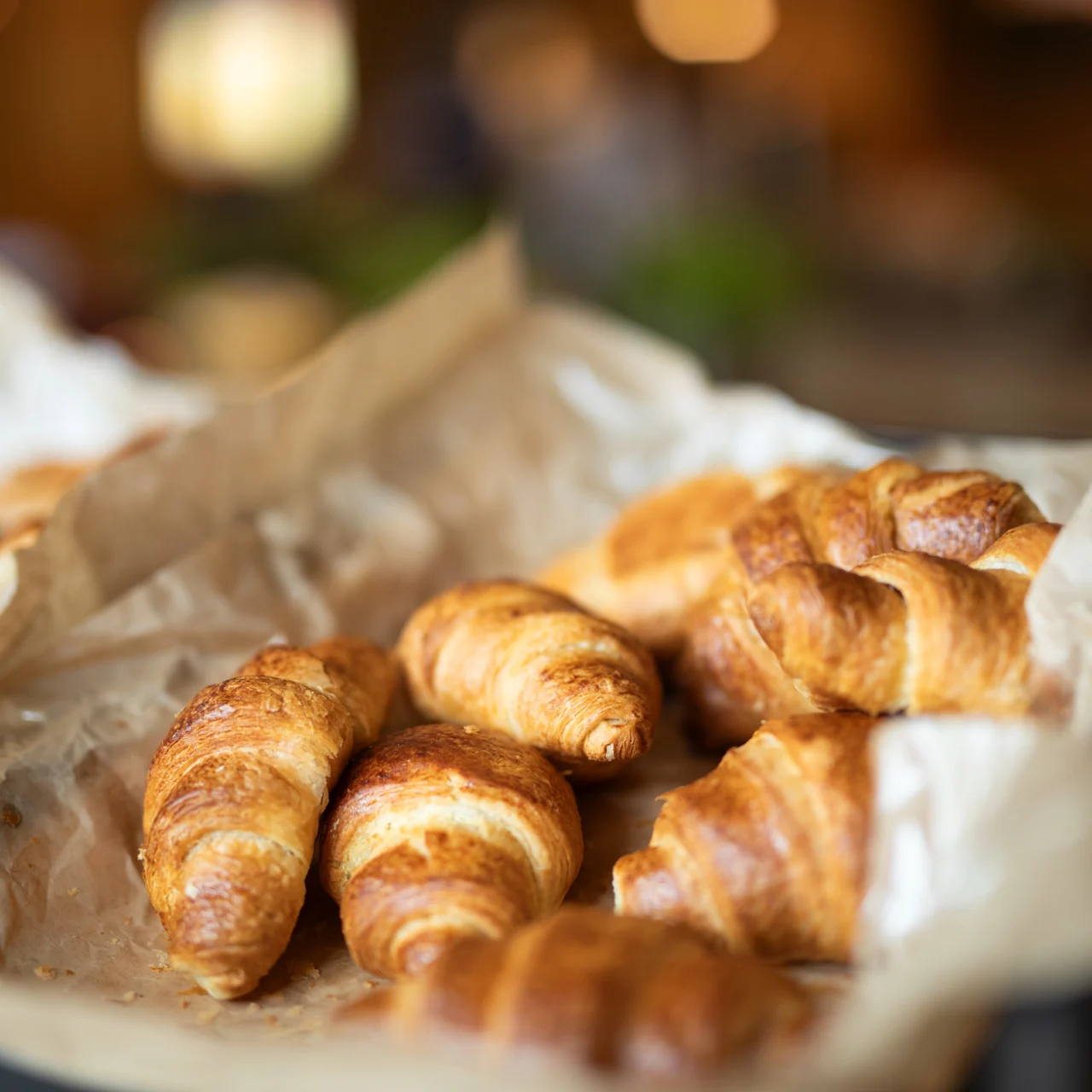 Färska croissanter ligger på skrynkligt bakplåtspapper, placerade i en korg i en mysig kafémiljö. Varm belysning förstärker bakverkens gyllenbruna färg. Ingen text synlig.