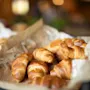 Nystekte croissanter hviler på krøllet bakepapir, plassert i en kurv i en koselig kaféinnstilling. Varm belysning fremhever de gyldenbrune fargetonene til bakverkene. Ingen tekst er synlig.