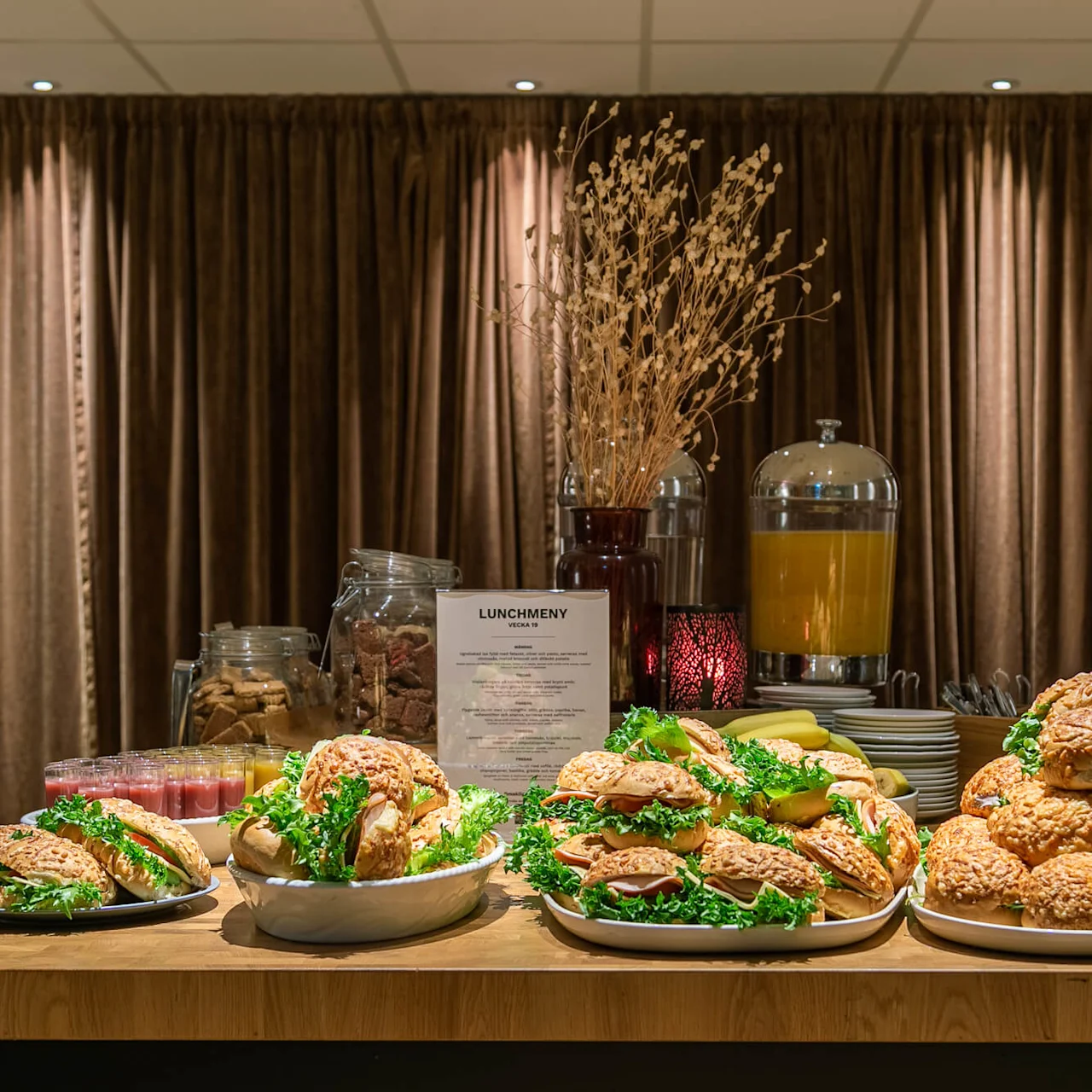 Sandwicher er arrangeret på fade med salat foran et brunt gardin. En glasbeholder med småkager, juice dispenser og en tørret plante pynter baggrunden. Tekst: "LUNCHMENY VECKA 47".
