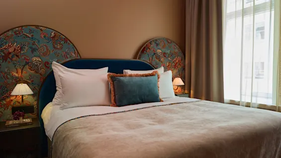 A neatly made double bed with patterned headboard, adorned with decorative pillows and side tables with lamps, stands in a softly lit room with beige walls and large curtained windows.