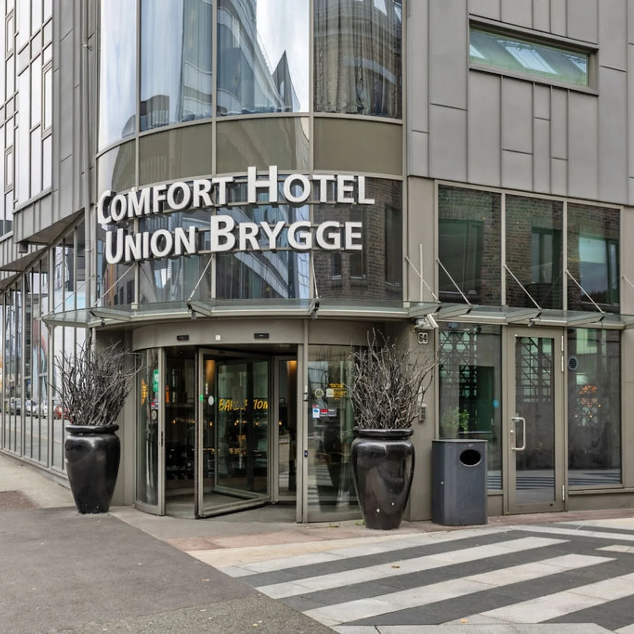 A modern hotel entrance features large glass doors beneath the sign "COMFORT HOTEL UNION BRYGGE." Two tall planters flank the entrance in an urban setting with a crosswalk nearby.