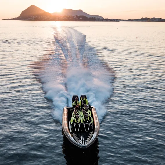 En rask RIB-båt med passasjerer som kjører i stor fart på fjorden i solnedgang.