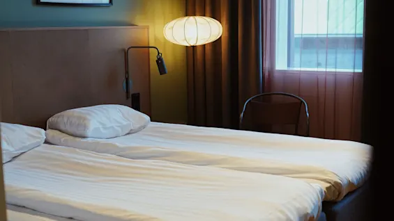 En prydligt bäddad säng med vita lakan står i ett mysigt rum, upplyst av en hängande lampa bredvid ett fönster med gardiner och en stol. 