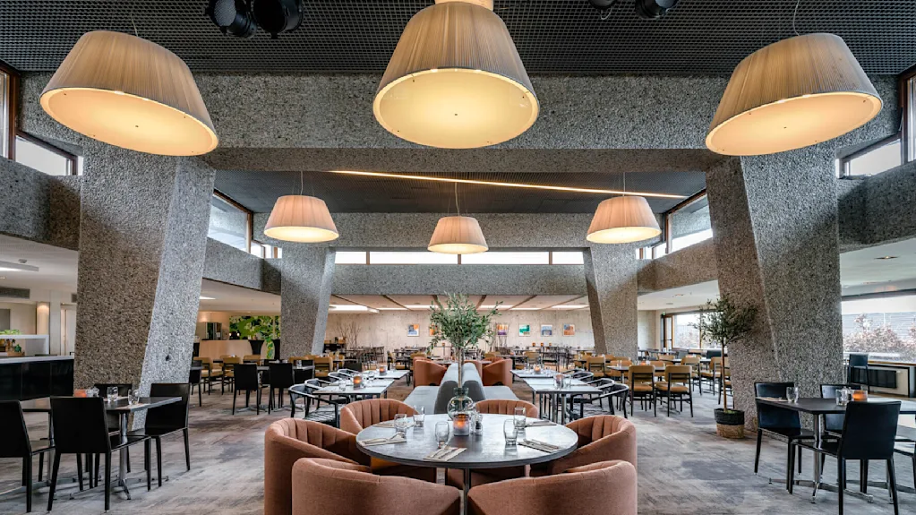 En restaurantinteriør har plysjstoler rundt runde bord. Store, moderne taklamper henger ovenfor og lyser opp det romslige området med polerte gulv og moderne dekor.