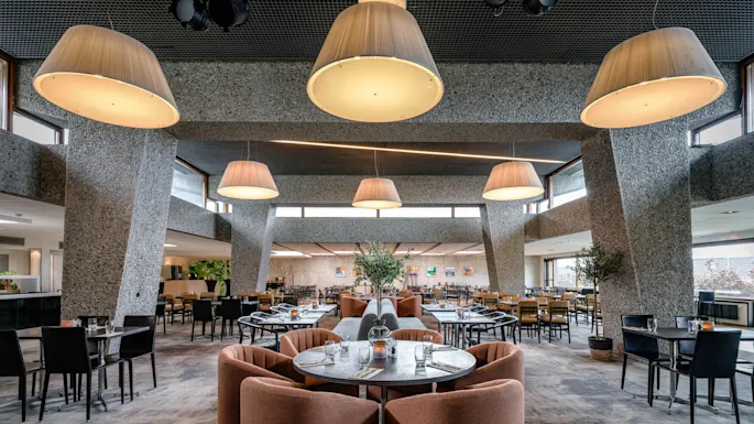 A restaurant interior features plush chairs around circular tables. Large, modern ceiling lamps hang above, illuminating the spacious area with polished floors and contemporary decor. 