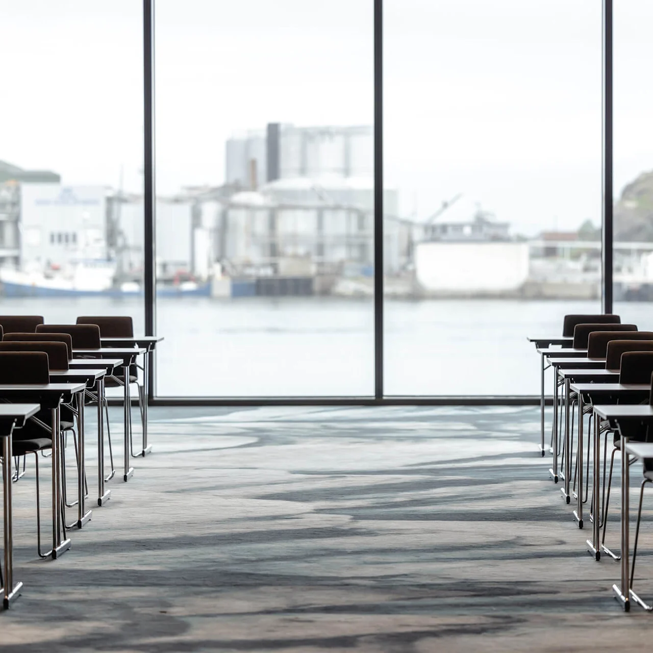 Rows of empty conference tables and chairs face large windows overlooking an industrial waterfront, creating a spacious meeting area with a scenic view.