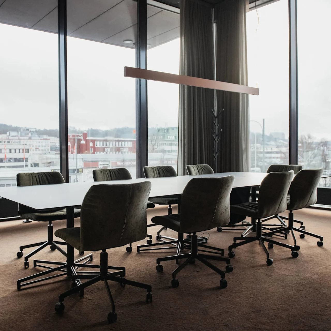 Ett stort konferensbord omges av gröna snurrstolar i ett rum med fönster som vetter mot byggnader. En taklampa hänger ovanför. Utanför står det "MIO."