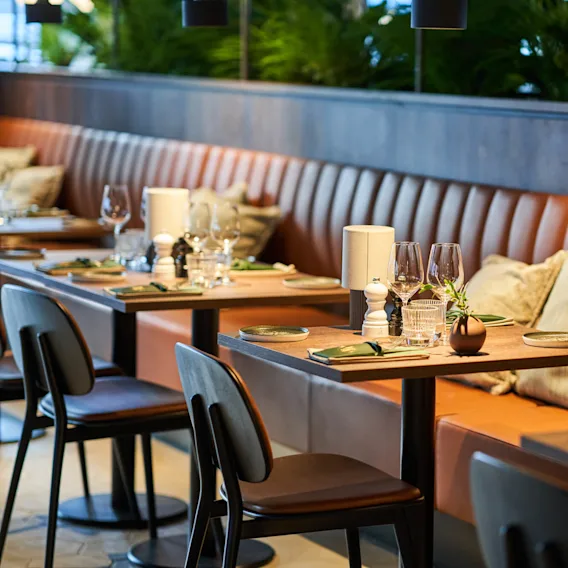Bord dukade med vinglas och servetter på en träyta; mjuka bruna läderstolar och grönska skapar en mysig restaurangmiljö.