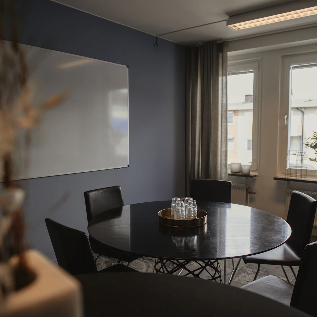 Round table with chairs holding a tray of glasses, set in a meeting room with a whiteboard, flip chart, and large windows showing an urban setting.