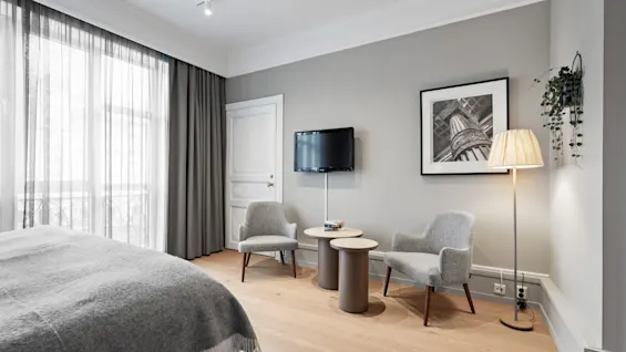 A stylish hotel room with a large bed, a comfortable seating area, a wall-mounted television, and natural light from a window.