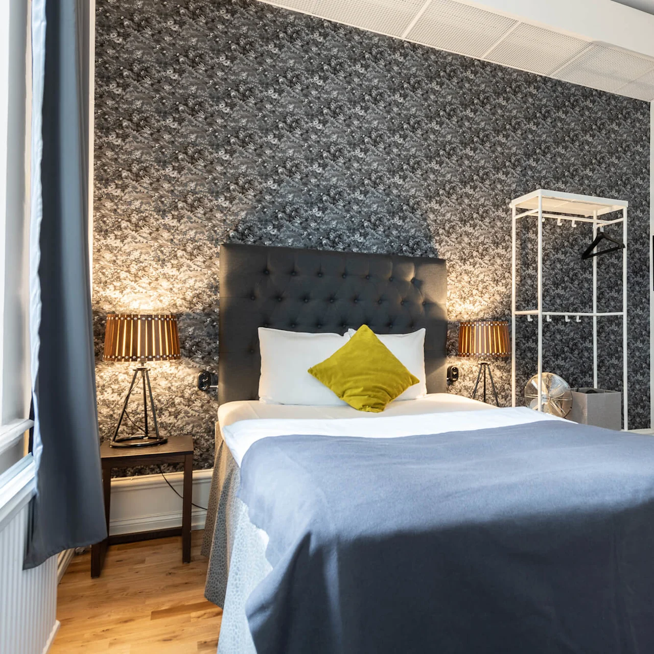 A neatly made bed with a dark headrest and a decorative pillow in a bedroom with elegant decor and luminous lamps.