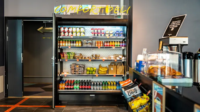 A refrigerated display shelves snacks, drinks, and fruits. Above, a neon sign reads “COMFORT DELI.” Nearby, a counter has chips and baked goods. A door labeled "403-416" opens to a hallway.
