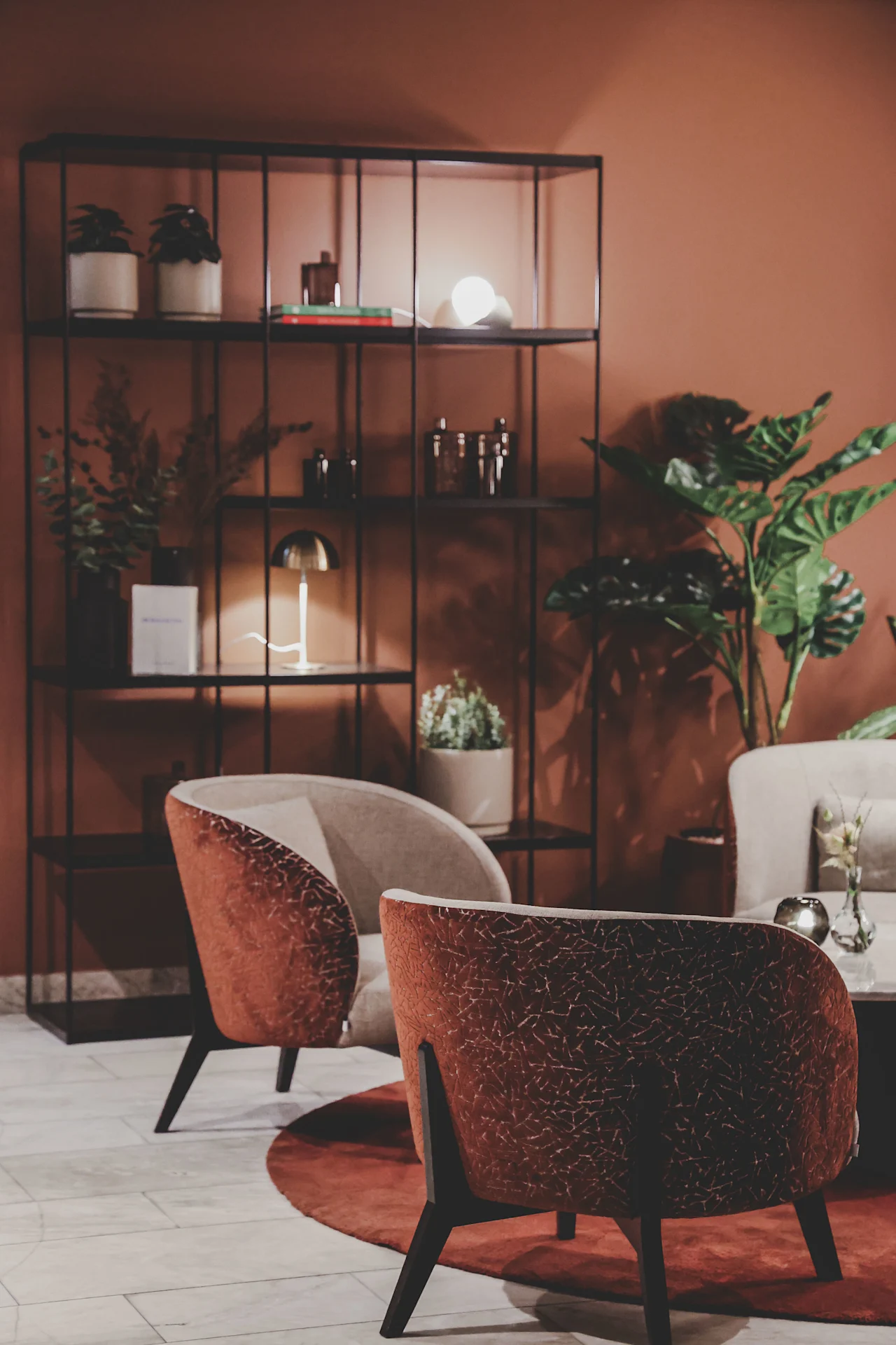 Two cushioned chairs sit on a circular rug, facing a metal shelf with decorative items, plants, and a lamp. A large plant adds greenery to the warm, ambient room.