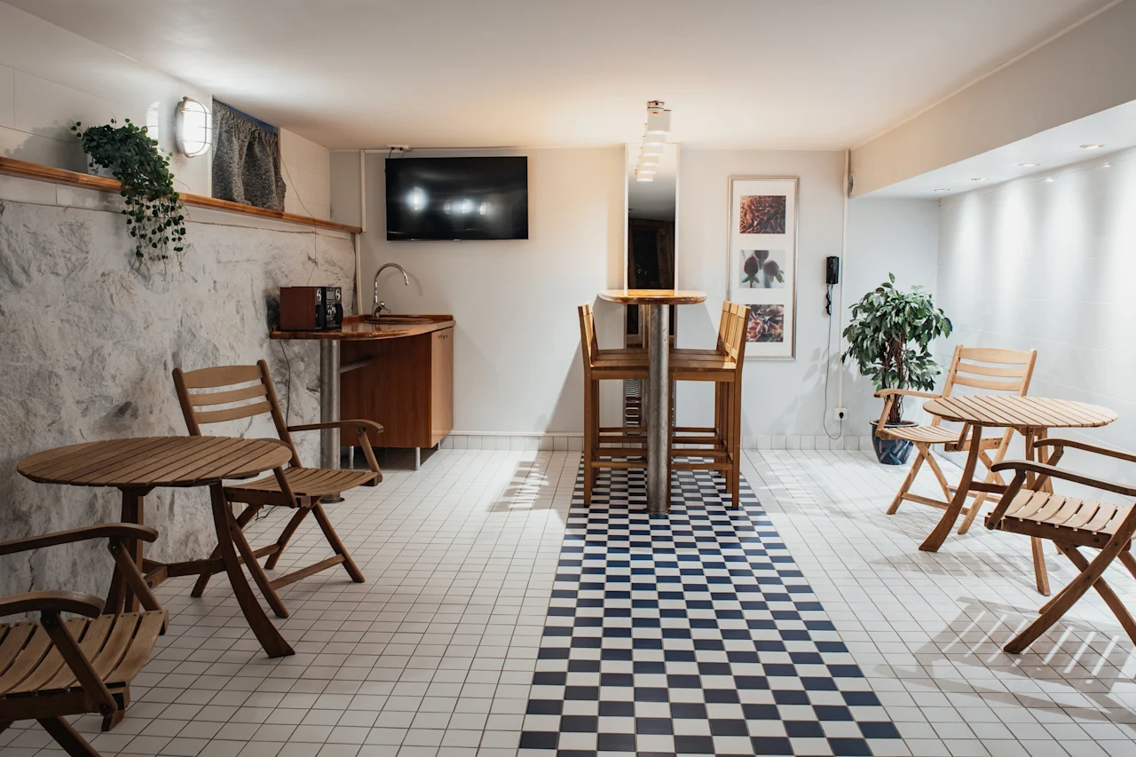 Home Hotel Kung Oscar: A cozy lounge area with wooden tables and chairs, a small kitchenette, and a checkered floor.