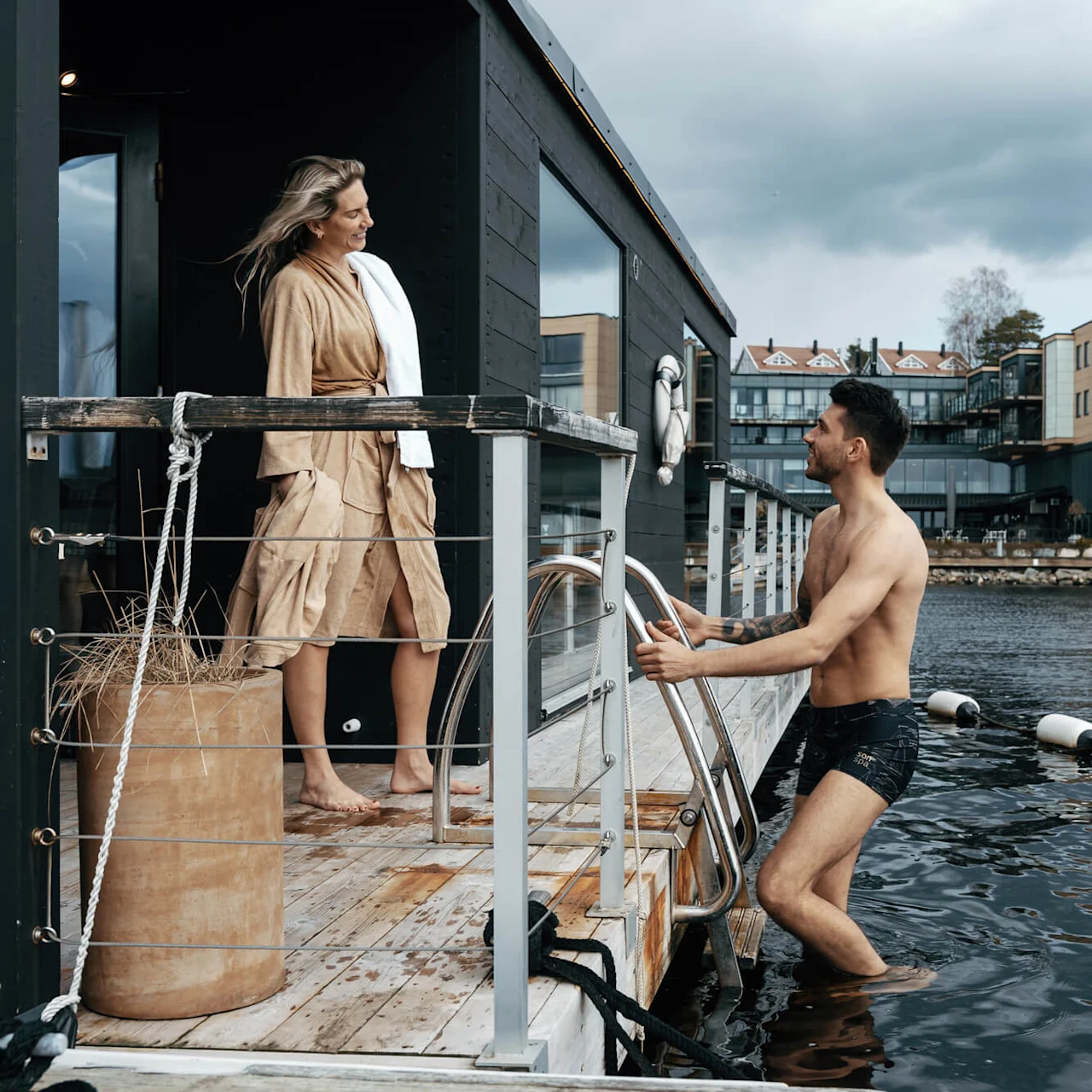 En skjorteløs mann klatrer opp en stige fra vannet til en trebrygge hvor en kvinne i badekåpe står, med bygninger synlige over vannet.