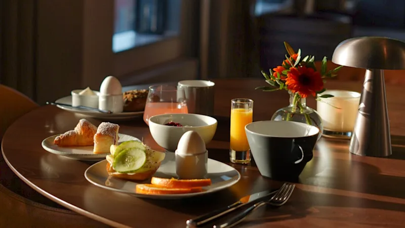 Hyvin valaistu ruokapöytä, jossa aamiaistuotteita kuten munia, kroissantteja, hedelmiä, mehua ja kahvia. Ympäröivät tuolit, kukat koristavat ja ikkuna valaisee huonetta luonnonvalolla.
