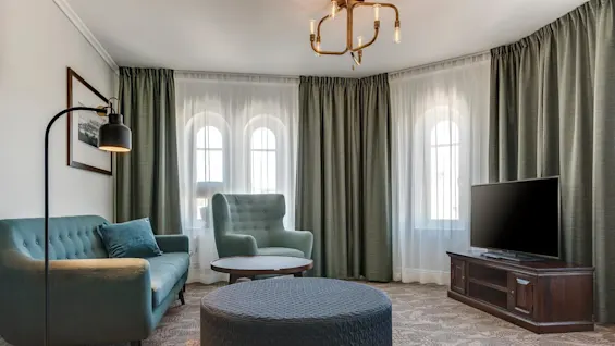 Elegant stue med trukket turkis sofa, matchende lenestol og rund puff, på mønstret teppe og under moderne lysekrone, flankert av vinduer med gardiner og TV.