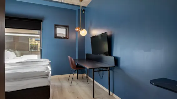 Et moderne hotellrom med blå vegger, en komfortabel seng, et skrivebord med TV og et vindu som gir naturlig lys.