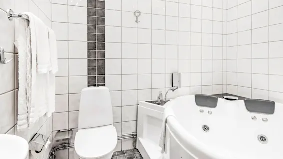 En hvit toalett står mot flislagte baderomsvegger med et håndkle på en hylle ved siden av, ved siden av et stort boblebad i en moderne, stilren setting.