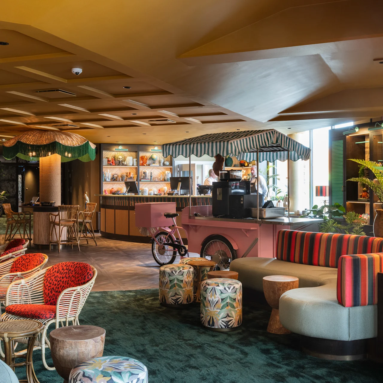 A seating area with wicker chairs and colorful cushions surrounds low tables. Nearby, a pink café counter with a canopy serves drinks. The setting has warm lighting and eclectic decor.