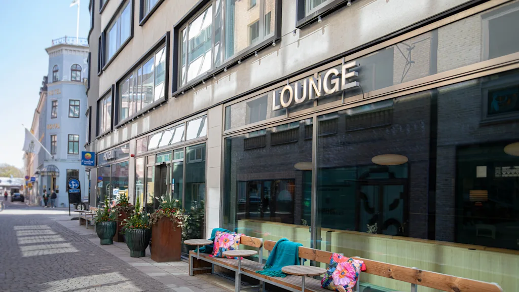 Et indbydende udendørs loungeområde med bænke og planter foran en bygning med store vinduer. Skiltet siger LOUNGE.