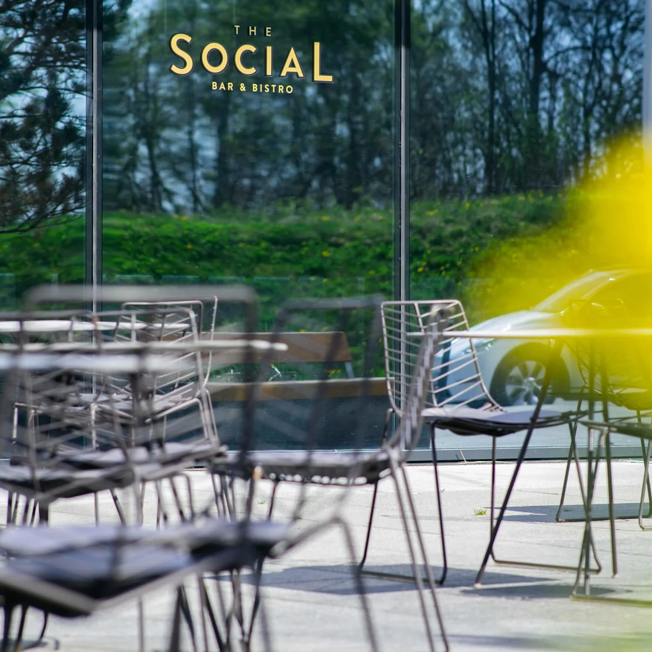 Tomme metallstoler og bord står på en terrasse. Reflekterende glass viser trær. Gult abstrakt uskarphet i forgrunnen. Tekst på glasset: "THE SOCIAL BAR & BISTRO".