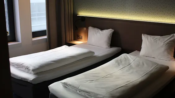 Two neatly made single beds with white linens are positioned side by side in a dimly lit hotel room featuring a patterned wall and a window to the left.