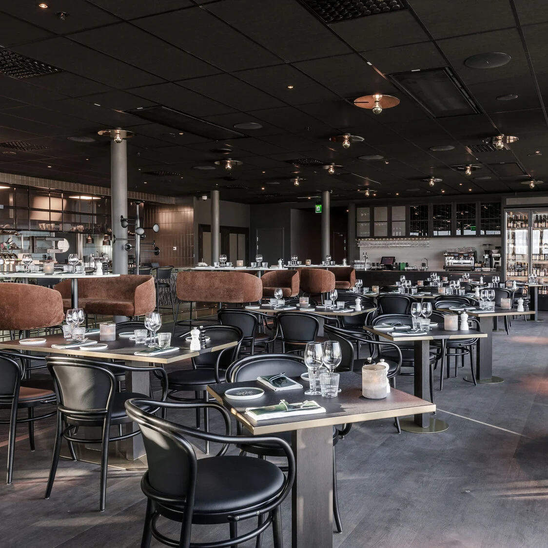 En rummelig, elegant dækket restaurantområde med runde borde og pænt arrangeret bordservice. Plydstole omgiver bordene, og store vinduer tilbyder udsigt. Hængende lamper ovenfor.