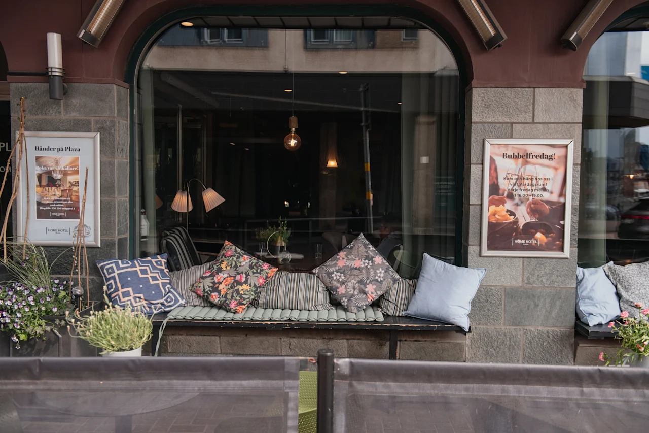 Kutsuva ulkoistuinalue, jossa on kuvioituja tyynyjä penkillä, suurten ikkunoiden ja kahden mainosjulisteen kehystämänä: Händer på Plaza ja Bubbelifredag!