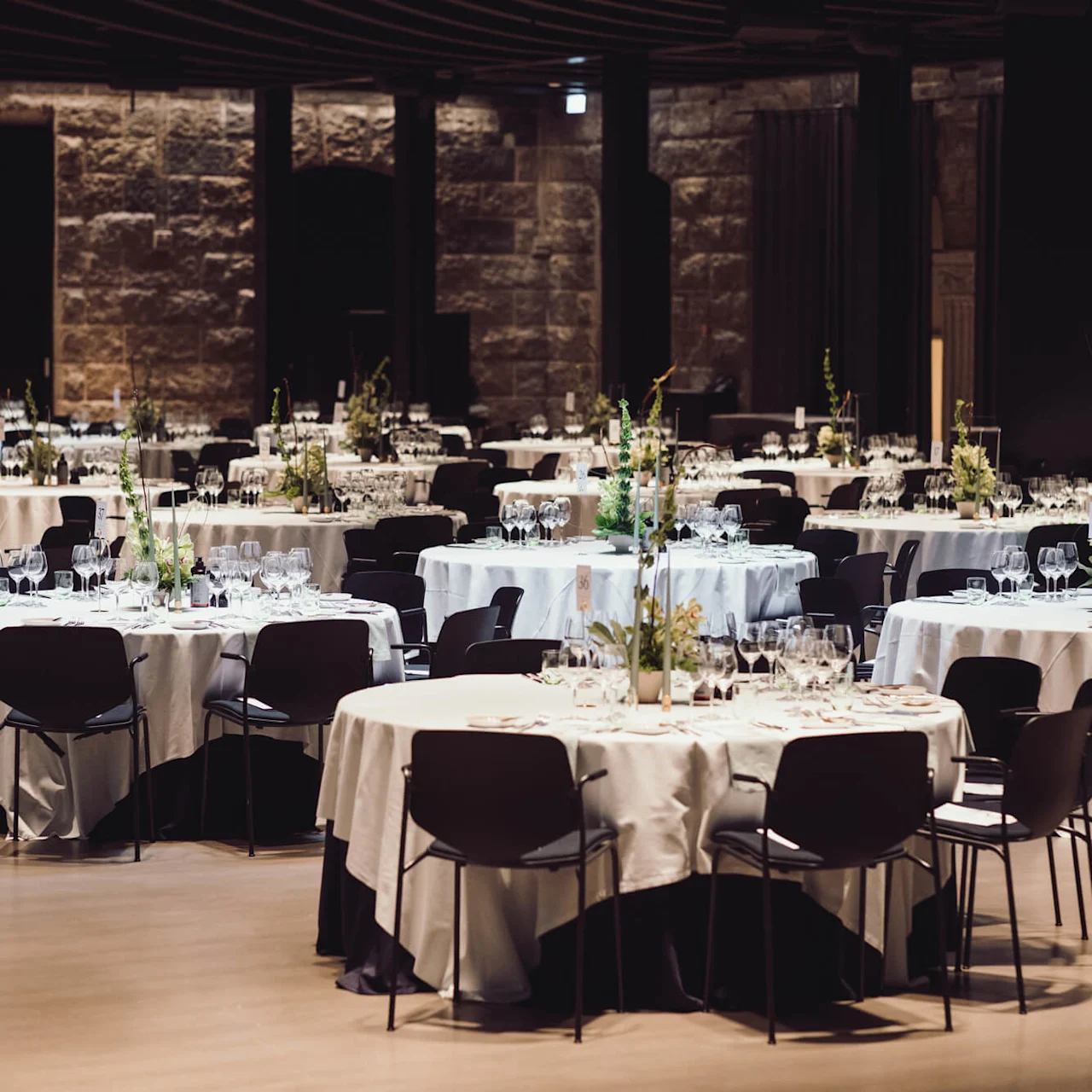Tables are elegantly set with white tablecloths, glassware, and floral centerpieces. Black chairs surround them in a spacious, dimly lit hall with stone walls.