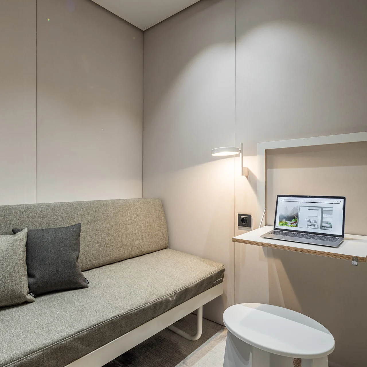 En grå soffa med två kuddar står i ett minimalistiskt rum. En bärbar dator på en vägghylla visar en webbsida. En modern lampa lyser upp området.