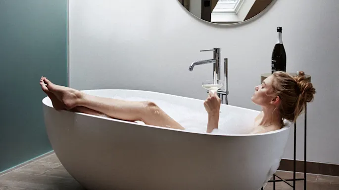 Woman relaxing in a bubble bath, holding a wine glass, in a modern bathroom with a round mirror and champagne bottle on a stand.